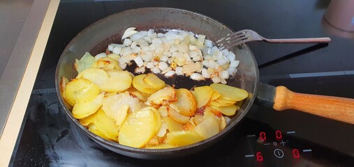 Thinly sliced potatoes and onions are fried in a cast iron pan. The pan is on an induction hob. The fork is visible in the frame, which is used to stir the potatoes