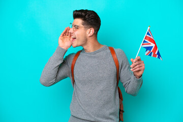 Young caucasian man holding an United Kingdom flag isolated on blue background shouting with mouth wide open to the side