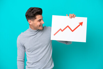 Young handsome caucasian man isolated on blue bakcground holding a sign with a growing statistics arrow symbol with happy expression