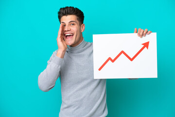 Young handsome caucasian man isolated on blue bakcground holding a sign with a growing statistics arrow symbol and shouting