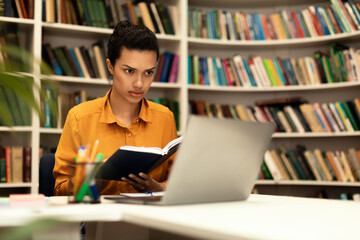 Focused mixed race lady reading book and looking at laptop, studying online, sitting in library, free space