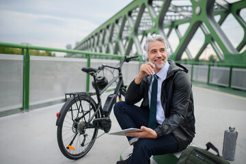 Businessman with bike sitting on bridge, using tablet and listening to music. Commuting and...