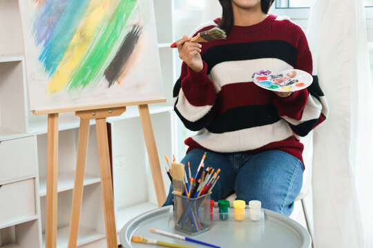 The art concept, Asian female artist in sweater holding paint brush and color palette in art studio