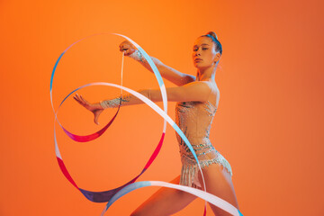 Young sportive girl, rhythmic gymnastics artist dancing isolated on orange color background. Concept of sport, action, aspiration, education, active lifestyle