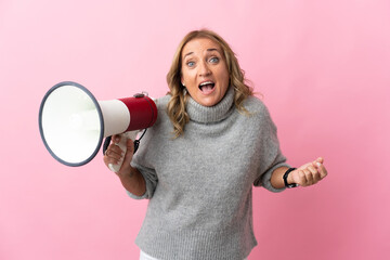 Middle aged blonde woman over isolated pink background holding a megaphone and with surprise...