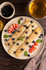Russian thin pancakes, rolled into tubes, on a beige plate on a dark background, strewn with blueberries with strawberries and mint, breakfast