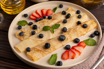 Russian thin pancakes, rolled into tubes, on a beige plate on a dark background, strewn with blueberries with strawberries and mint, breakfast