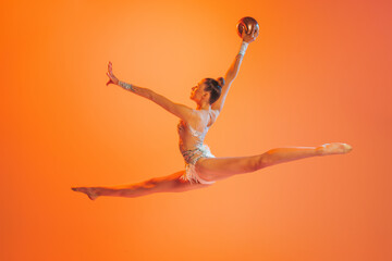 One professional rhythmic gymnastics artist training with golden color ball isolated on orange...