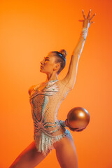 One professional rhythmic gymnastics artist training with golden color ball isolated on orange...