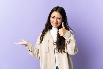 Young caucasian woman isolated on purple background holding copyspace imaginary on the palm to insert an ad and with thumbs up