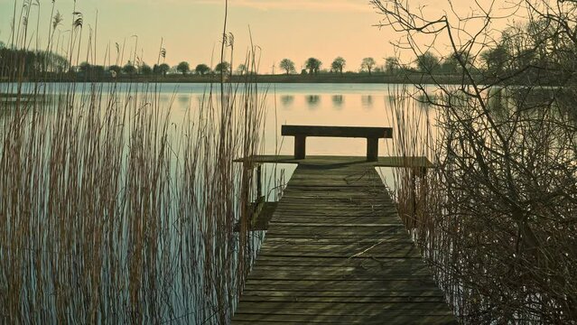 A wharf stretches out over Lake Assens between reeds and bushes illuminated by the warm light of the spring sunset. Assens, Denmark, Europe
