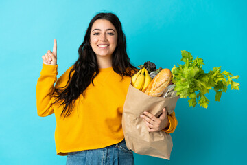 Young woman holding a grocery shopping bag isolated on blue background pointing up a great idea