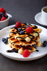 Viennese or Belgian waffles with fresh berries (raspberries and blueberries) on a white plate and a cup of coffee. Traditional dessert. Close-up, grey background.