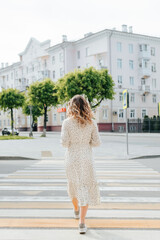 Stylish young woman with curly hairstyle crossing road outdoors, back view. Single slender woman wearing white summer dress walks along pedestrian crossing on street in city