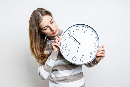 Young woman looks at wall white clock in her hands on a light background.