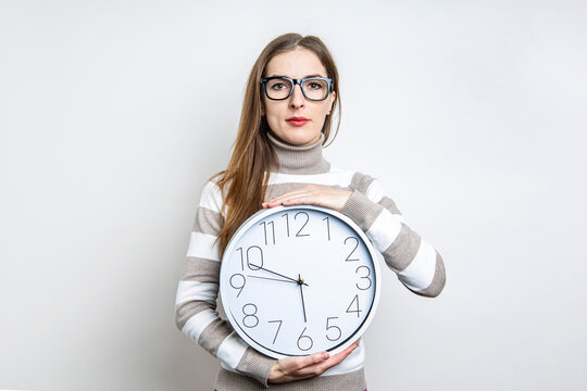 Young woman in glasses holding a wall clock on a light background.