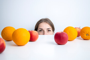 Fototapeta na wymiar Young woman peeks out from under a table on which fruits lie against a white wall