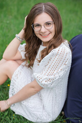 Smiling brunette woman in eyeglasses sitting back to back with unrecognizable man grass in park and looking at camera