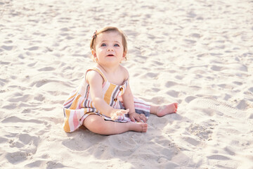 happy baby girl sitting on the beach on sand wearing striped summer dress on sunny day