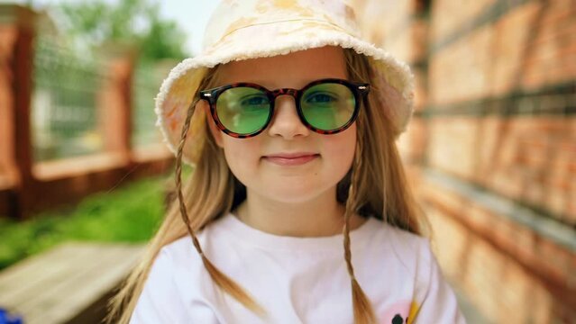 Portrait little stylish kid girl in summer sunglasses and a panama on city street. Child smiles and looks at camera. 