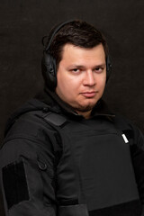 Obraz premium Portrait of a man 25-30 years old in a bulletproof vest and tactical headphones for shooting on a black background.