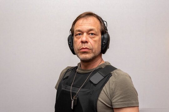 An Elderly Man 45-50 Years Old In A Bulletproof Vest And Tactical Headphones For Shooting On A Light Background.