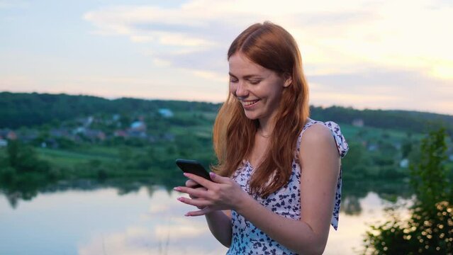 Attractive girl surfing internet, social media stands near lake play her phone. Pretty smiling ginger woman with freckles using smartphone on nature background, sending picture having fun outdoors.