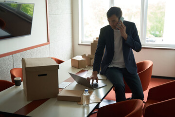 Man among unwrapped boxes using laptop during phone talk