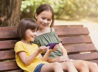 Foto op Plexiglas Twee gelukkige meisjes kijken naar hun telefoon. Mooie kinderen die internetten in het park © Albert Ziganshin
