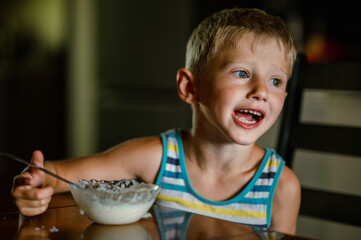 a cute boy with blond hair in a striped T-shirt is having breakfast at home in the kitchen, eating porridge and drinking milk from a glass glass