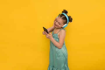 Fototapeta na wymiar Cheerful positive girl holds a smartphone, has a modern blue headset, enjoys stereo sound melody or dances to music on a yellow background