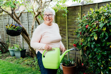 Portrait of happy, smiling senior, mature woman watering flowers with watering can in the garden in...