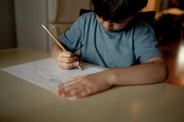 a five-year-old boy in a blue T-shirt is sitting at home at the table and drawing with pencils, the child is passionate about drawing, hobby