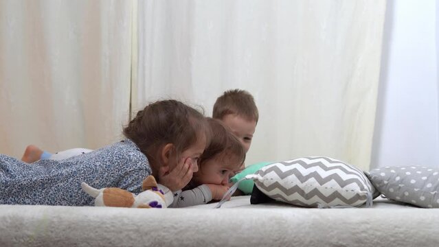 Smiling Preschool Toddler Children In Pajamas Watching Cartoon on Smartphone on Bed. Siblings Little Twins Boy and Girl Have Fun. Happy Kids On Quarantine At Home. Friendship, Family, Education