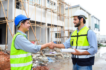 Construction engineer and architect with blueprint shaking hands while standing on construction site. Home building concept.