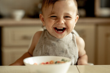 cute smiling baby eats colorful cereal flakes for breakfast. The child is happy, he is sincerely...