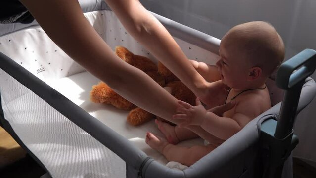 Close-up Cute Kid 10 Month Newborn Boy Crying in White Crib in morning. Sad Child Face biting gnaws everything. teething, gum pain. Childhood, Motherhood, Family, Infant. mother takes baby in her arms