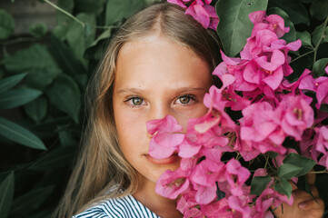 portrait face of candid beautiful little kid girl of eight years old with green brown eyes on background of green plants and pink flowers during a summer vacation travel. gen z mental health concept