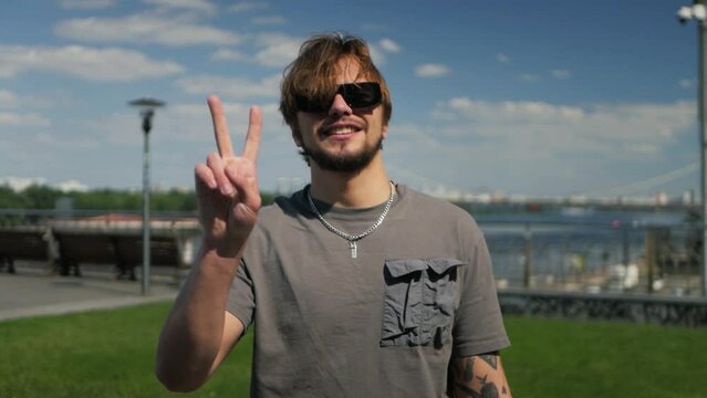 Cheerful smiling caucasian dude with tattoed arm showing the peace sign outdoors. Good-looking man lifting arm and performing V gesture on bright sunny day. Male model signaling for peace in slow mo
