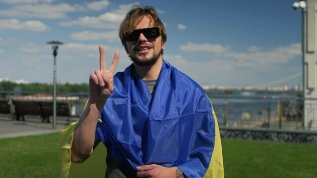 Beaming ukrainian man wearing national flag showing the peace gesture on sunny day. Young stylish dude with blue and yellow flag on shoulders performing  victory sign and smiling spreading positivity
