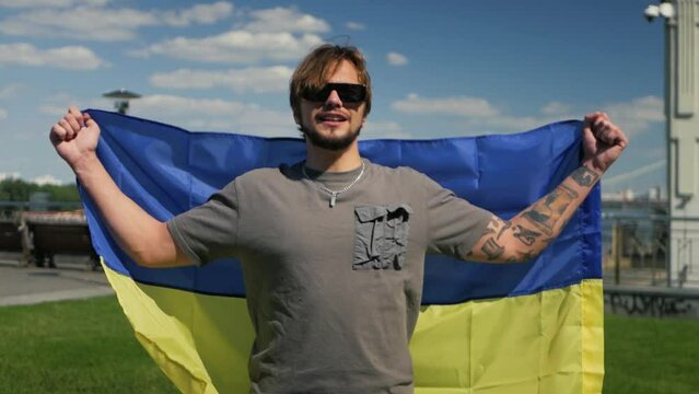 Modern caucasian man showing support for Ukraine by wearing national flag and celebrating. Slow-mo shot of stylish dude in sunglasses lifting blue and yellow flag above head and commemorating peace
