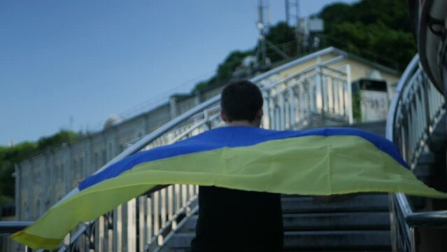 Ukrainian man wearing blue and yellow flag moving toward his goal with resilience despite everything. Slow motion shot of caucasian man running up the stairs with ukrainian banner behind his back
