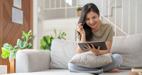 Beautiful attractive young Asian woman sitting on gray sofa and using digital tablet at living room in the morning. - Powered by Adobe