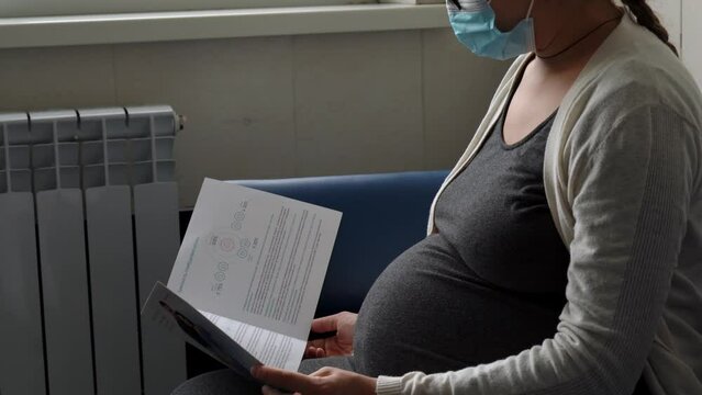 Young Pregnant Woman In Medicine Protective Mask Reading On Couch Waiting For Appointment Medical Control On Hospital. Visit Gynecologist Doctor At Clinic. Examine Expectant Belly Healthcare Check Up