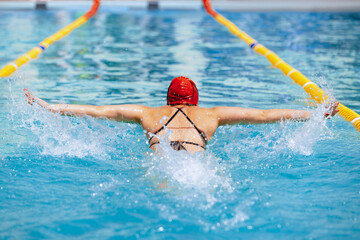 Back view of sportive woman, swimmer in swimming cap and goggles practicing at public swimming-pool, outdoors. Sport, power, energy, style, hobby concept.