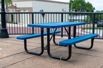 A vibrant blue and black empty metal picnic table on a ceramic patio of a beer garden. There's a white concrete shop in the background with trees and a parking area. 
