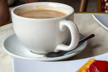 A white porcelain cup and saucer with a small teaspoon on the saucer. The mug is filled with hot...