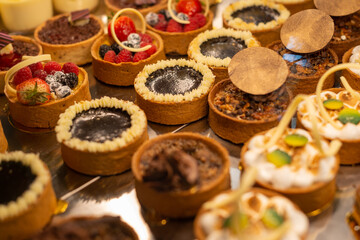 Rows of sweet pastries in a glass showcase at a patisserie. There are small round baked cakes, chocolate mousse, pastries, biscuits, berry tarts, and fruit snacks on display for sale. 