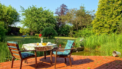 romantischer Frühstücksplatz am Gartenteich mit Holzbrücke neben roter Terrasse und grünem Garten