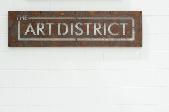 A Distressed Metal Sign With The Text Art District Cutout. The Sign Is Hanging On A White Brick Wall. The Signage Is For A Public Space For Galleries, Theatres And Modern Performances.  